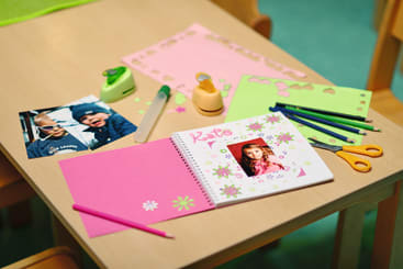 Eine Tischplatte mit Scrapbook-Zubehör, darunter farbiges Papier, Scheren, Klebstoff, Stifte, ein grüner Locher, Fotos und ein aufgeschlagenes Notizbuch, das mit Blumen und einem Foto mit der Aufschrift Kate verziert ist.