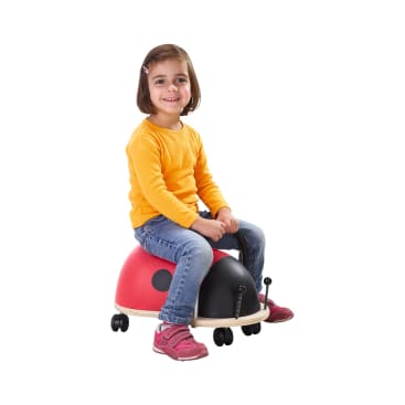 Ein kleines Kind in einem gelben langärmeligen Hemd, blauen Jeans und rosa Schuhen sitzt lächelnd auf einem fahrbaren Spielzeug in Form eines Marienkäfers und hält sich an einem schwarzen Griff fest.