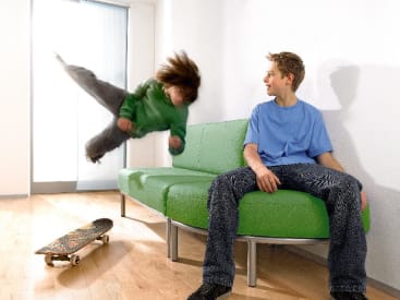 Eine Person in einem grünen Sweatshirt ist in der Luft gefangen und fällt auf eine grüne Couch, während eine andere Person in einem blauen Shirt auf der Couch sitzt und sie anschaut. Ein Skateboard liegt auf dem Holzboden in der Nähe.