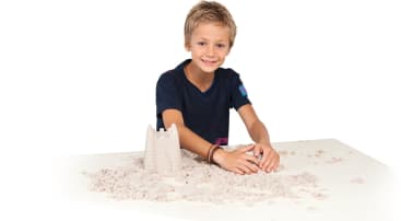 Ein kleiner Junge sitzt an einem weißen Tisch und formt lächelnd Sand mit seinen Händen. Vor ihm ist eine kleine Sandburg gebaut, und loser Sand ist auf dem Tisch verteilt. Der Hintergrund ist einfarbig weiß.