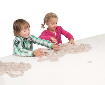 Zwei kleine Kinder, ein Junge und ein Mädchen, sitzen an einem weißen Tisch und spielen mit hellem Sand. Der Junge trägt ein grün kariertes Hemd, das Mädchen eine rosa Strickjacke. Beide sind auf den Sand vor ihnen konzentriert.