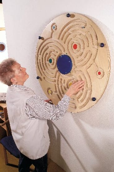 Eine Person steht vor einem an der Wand befestigten runden Holzlabyrinth und bewegt ihre Hand entlang der Rillen. Das Labyrinth weist mehrere farbige Kreise und Pfade auf, und die Person scheint sich auf ihre Tätigkeit zu konzentrieren.