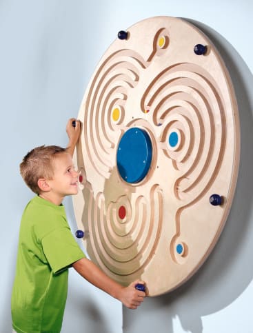 Ein Junge in einem grünen Hemd steht neben einem großen, kreisförmigen Holzlabyrinth, das an einer Wand befestigt ist, und dreht einen Knopf, um Murmeln durch die Rillen zu bewegen. Das Labyrinth hat farbige Löcher und eine blaue Scheibe in der Mitte.