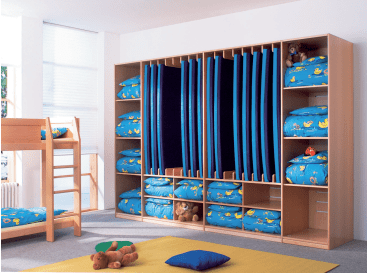 Ein Kinderzimmer mit einer Reihe von Holzregalen, in denen mehrere blaue Matratzen und Bettzeug mit Cartoon-Enten aufbewahrt werden. Auf der linken Seite steht ein Etagenbett, und auf dem gelben Teppich auf dem Boden liegen Spielzeug und ein Kissen.