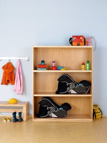 In einem Holzregal stehen zwei schwarze Autositze für Kleinkinder, Flaschen und Kindersachen. In der Nähe stehen eine Bank mit Schuhen, ein Kleiderständer mit Jacken, ein kleiner Rucksack und eine gelbe Tasche auf dem Boden.