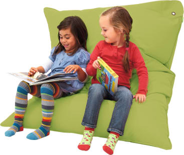 Zwei junge Mädchen sitzen auf einem großen grünen Bodenkissen. Ein Mädchen liest ein Buch vor, während das andere ein geschlossenes Buch hält und zuhört. Beide Mädchen tragen bunte Kleidung und Socken.