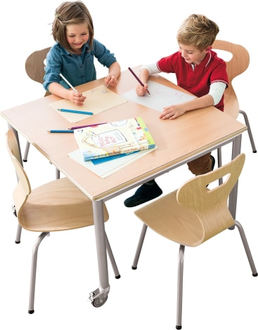 Zwei Kinder, ein Mädchen und ein Junge, sitzen an einem quadratischen Holztisch und zeichnen mit Buntstiften. Auf dem Tisch befinden sich zwei unbesetzte Stühle und einige Papiere und Bücher.