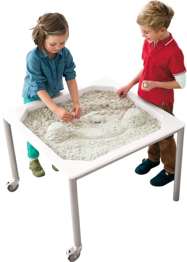 Zwei Kinder stehen an einem weißen Tisch mit einem erhöhten Rand und spielen mit hellem Sand. Der Sand ist zu Formen geformt, und der Tisch hat Räder an seinen Beinen. Der Hintergrund ist einfarbig weiß.
