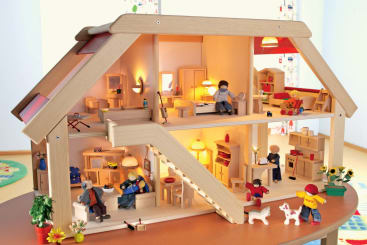 Ein Puppenhaus aus Holz mit zwei Etagen, möblierten Zimmern und mehreren kleinen Figuren, die eine Familie bei verschiedenen Aktivitäten darstellen. Das Haus ist hell erleuchtet, in der Nähe stehen Pflanzen und Spielzeug.