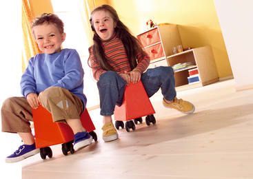 Zwei kleine Kinder sitzen auf kleinen Rollstühlen, lächeln und spielen in einem hell erleuchteten Raum mit Holzfußboden und Regalen im Hintergrund.