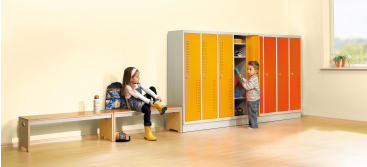 Zwei Kinder befinden sich in einem hell erleuchteten Raum mit Schließfächern. Ein Kind sitzt auf einer Bank und zieht sich die Schuhe an, während das andere an einem offenen Spind steht und hineingreift. Ein Fenster und ein Rucksack sind ebenfalls zu sehen.