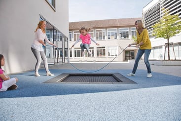 Ein Kind springt auf einem eingebauten Trampolin in einem Innenhof, während zwei Erwachsene ein Seil halten, über das es springen kann. Ein anderes Kind sitzt in der Nähe und schaut zu. Der Bereich hat einen blauen Gummibodenbelag und ist von Gebäuden umgeben.