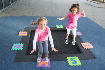 Zwei Mädchen spielen draußen auf einem eingebauten Trampolin, das von Schaumstoffpuzzleteilen mit großen Buchstaben umgeben ist. Beide Mädchen sind mitten im Sprung, und die Umgebung umfasst einen gepflasterten Boden und einen blauen Gummispielbereich.