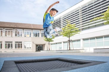 Ein Junge in blauem Hemd und Jeans springt energisch auf einem viereckigen Trampolin, das in den Boden eines Innenhofs eingelassen ist, mit modernen Schulgebäuden und kleinen Bäumen im Hintergrund.