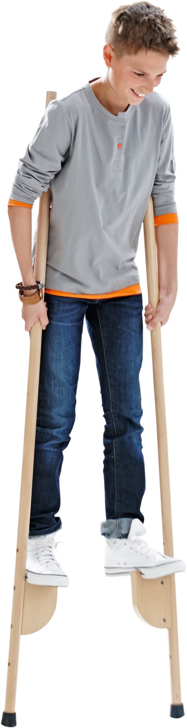 Ein Jugendlicher in einem grauen, langärmeligen Hemd und blauen Jeans steht auf Holzstelzen. Er lächelt und schaut nach unten, während er auf den Stelzen balanciert. Der Hintergrund ist weiß.