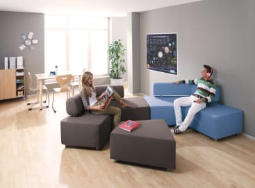 Zwei junge Menschen sitzen auf modernen, modularen Sofas in einem hellen Raum mit Holzboden. Einer liest eine Zeitschrift, während der andere Kopfhörer trägt. Im Hintergrund stehen ein Schreibtisch, Stühle, Bücherregale und eine Pflanze.