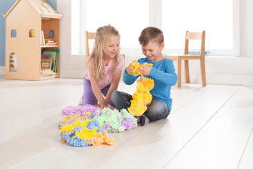 Zwei Kinder sitzen auf einem weißen Boden und spielen mit bunter Schaumstoffknete, die sie zu langen, strukturierten Stücken formen. Im Hintergrund stehen ein hölzernes Puppenhaus und Stühle. Der Raum ist hell und aufgeräumt.