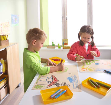 Ein Junge und ein Mädchen sitzen an einem weißen Tisch und zeichnen mit Buntstiften. Der Junge skizziert ein Haus, während das Mädchen mit Grün malt. Helle Tabletts mit Stiften stehen auf dem Tisch, und Sonnenlicht fällt durch ein Fenster hinter ihnen.