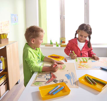 Zwei kleine Kinder sitzen an einem weißen Tisch in einem hell erleuchteten Raum und malen bunte Bilder mit Buntstiften auf große Papierbögen. Die Malutensilien sind in gelben Fächern auf dem Tisch angeordnet.