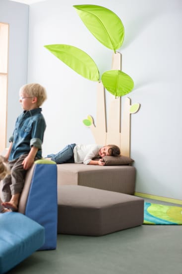Ein kleines Kind liegt auf einer grauen, gepolsterten Plattform unter großen Blattdekorationen an der Wand, während ein anderes Kind in der Nähe steht. Bunte Spielmatten und weiche Formen sind in dem hellen Spielzimmer zu sehen.