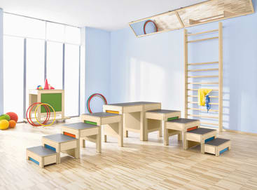 Ein hell erleuchtetes Spielzimmer mit einer hölzernen Klettertreppe, einer an der Wand befestigten Leiter, bunten Bällen, Reifen und Turngeräten, die auf einem hellen Holzboden vor blauen Wänden angeordnet sind.
