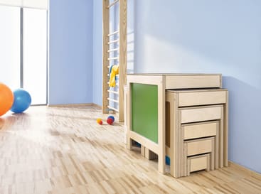Ein heller Raum mit hellem Holzfußboden, einer Wandleiter, einigen bunten Bällen und einem Satz stapelbarer Holztische mit einer grünen Platte an der Seite, vor einer blauen Wand. Sonnenlicht strömt durch ein Fenster herein.