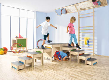 Vier Kinder spielen in einem hell erleuchteten Raum mit verschiedenen hölzernen Kletterstrukturen. Drei Kinder klettern und laufen auf den Stufen, während ein Kind darunter hindurchkrabbelt. Im Hintergrund sind bunte Bälle und Reifen zu sehen.