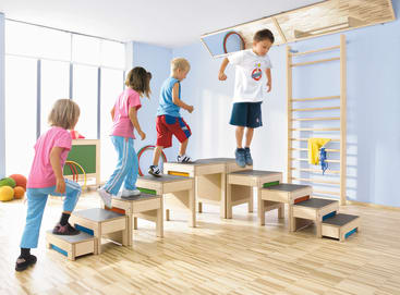Vier kleine Kinder spielen und klettern auf einem hölzernen Indoor-Spielgerät mit Stufen und Plattformen in einem hell erleuchteten Raum mit blauen Wänden, Holzfußboden und Turngeräten im Hintergrund.