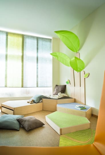 Ein helles Spielzimmer mit großen Fenstern, weichen Bodenkissen, niedrigen Holzpodesten mit grünen und grauen Platten, einem kleinen Bücherstapel und an der Wand angebrachten blattförmigen Dekorationen, die an einen Baum erinnern.