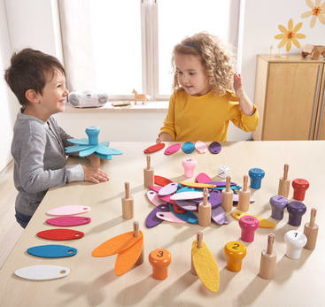 Zwei Kinder sitzen an einem Tisch und spielen mit einem bunten Zähl- und Stapelspielzeug aus nummerierten Holzstiften und Filzblättern. Der Tisch ist mit Spielzeugteilen bedeckt, und die Kinder scheinen mit ihrer Tätigkeit beschäftigt zu sein.