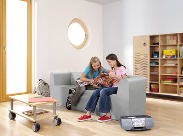 Zwei Mädchen sitzen auf einer hellgrünen Couch in einem Raum mit Holzboden und schauen sich gemeinsam Zeitschriften an. Ein Rucksack, ein Wagen mit Büchern und eine Stereoanlage stehen in der Nähe. Ein Bücherregal und ein rundes Fenster befinden sich im Hintergrund.