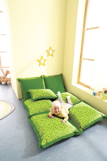 Ein kleines Kind liegt lächelnd auf einem provisorischen Bett aus grün gemusterten Bodenkissen in einem hellen Raum mit gelben Wänden, zwei sternförmigen Wandspiegeln und einem großen Fenster, durch das Sonnenlicht fällt.