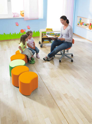 Eine Frau sitzt auf einem Stuhl und liest zwei kleinen Kindern vor, die auf orangefarbenen und grünen Hockern in einem hellen Klassenzimmer mit Holzfußboden und pädagogischen Dekorationen an den Wänden sitzen.