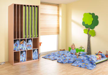 Zwei kleine Kinder liegen auf blauem Bettzeug mit Cartoon-Motiven auf Matten auf einem Holzboden. Hinter ihnen befindet sich ein hölzerner Schrank, in dem gerollte Matten und Taschen aufbewahrt werden. Das Zimmer hat ein Fenster mit Jalousien und eine Baumdekoration an der Wand.