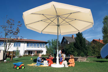 Mehrere kleine Kinder sitzen im Gras unter einem großen beigen Sonnenschirm in einem sonnigen Garten. In der Nähe sind ein Spielzeugauto und Bäume zu sehen. Im Hintergrund sind Häuser und ein weißes Gebäude mit großen Fenstern zu sehen.
