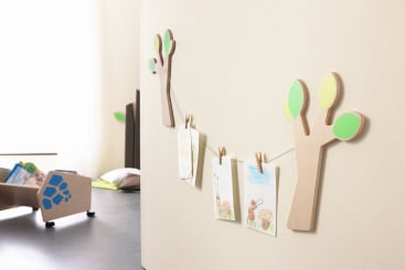An einer beigen Wand sind zwei baumförmige Holzdekorationen mit grünen Blättern angebracht, die durch eine Schnur verbunden sind, an der Kinderzeichnungen mit Wäscheklammern befestigt sind. Im Hintergrund befindet sich eine mit Büchern und Kissen gefüllte Kiste auf dem Boden.