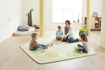 Eine Frau sitzt mit drei kleinen Kindern auf einem Teppich in einem hellen, geräumigen Zimmer. Die Kinder sitzen in einem Kreis. Der Raum hat einen Holzboden, helle Wände und Regale mit Spielzeug und Kissen.