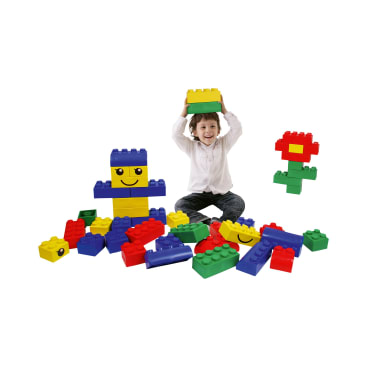 Ein Kind sitzt auf dem Boden, umgeben von großen, bunten Spielzeugbausteinen. Das Kind hält einen gelben Klotz über seinen Kopf und lächelt. Einige der Blöcke sind zu Formen mit Gesichtern und einer Blume zusammengesetzt.