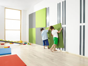 In einem hellen Raum mit Holzboden und Turnmatten befestigen zwei Kinder grüne gepolsterte Platten an einer weißen Wand mit vertikalen schwarzen Streifen. Verschiedene Spielgeräte sind auf dem Boden verstreut.