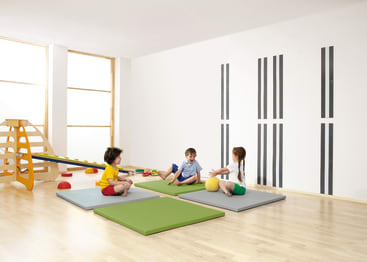 Drei Kinder sitzen auf quadratischen Matten in einem hellen, großen Raum mit Holzfußboden. Ein Kind hält einen gelben Ball. Im Hintergrund befinden sich ein Klettergerüst, ein Schwebebalken und einige Spielgeräte.