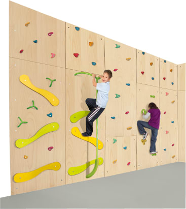 Zwei Kinder klettern an einer Indoor-Kletterwand aus Holz mit bunten Griffen und großen gelben und grünen Kletterelementen. Die Wand steht in einer Ecke eines Zimmers mit grauem Boden.