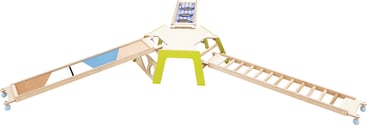 Ein Holzspielgerät mit drei Rampen, die von einer zentralen Plattform ausgehen. Eine Rampe ist mit blauen und beigen Paneelen versehen, eine andere mit Holzsprossen und die dritte mit einer Kletterwand aus Netz. Die Struktur ist auf Rädern aufgestellt.