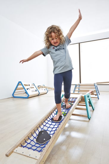 Ein kleines Kind mit lockigem Haar balanciert mit ausgestreckten Armen auf einer blauen Kletternetzbrücke in einem hellen, minimalistischen Spielzimmer mit Holzspielgeräten.