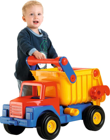 Ein kleines Kind steht hinter einem großen Spielzeug-Muldenkipper aus Plastik und hält sich an seinem gelben Behälter fest. Der Lkw ist rot, gelb und blau. Das Kind lächelt und trägt ein dunkelblaues Hemd und eine Jeans.