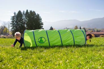 Zwei Kinder krabbeln draußen auf dem Gras und kommen an beiden Enden eines hellgrünen Spieltunnels heraus. Im Hintergrund sind Bäume, Häuser und Berge unter einem klaren blauen Himmel zu sehen.