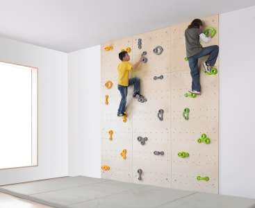 Zwei Kinder klettern an einer Indoor-Kletterwand aus Holz mit gelben, grauen und grünen Griffen. Die Wand ist in einem hellen, weißen Raum mit einem großen Fenster und einem hellen, gepolsterten Boden zur Sicherheit installiert.