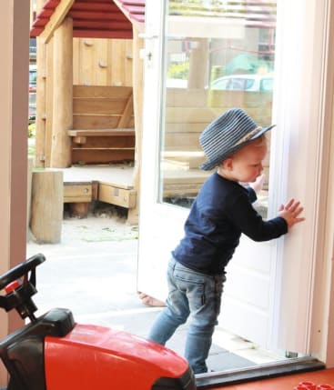 Ein kleines Kind mit blauer Mütze und Jeans schiebt eine Glastür auf, um nach draußen zu gehen, während im Hintergrund ein hölzernes Spielhaus und ein Sandkasten zu sehen sind. Ein rotes Spielzeugfahrzeug ist teilweise im Vordergrund zu sehen.