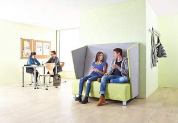 Zwei junge Leute sitzen auf einer grünen Bank mit hoher Lehne und unterhalten sich, während zwei andere an einem Tisch im Hintergrund in der Nähe eines Fensters und einer Pinnwand in einem hellgrünen Raum arbeiten. Mäntel und Taschen hängen an Haken an der Wand.