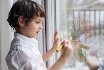 Ein kleines Kind mit kurzen braunen Haaren zeichnet mit einem gelben Marker auf ein Fenster. Das Kind trägt ein weißes Hemd und konzentriert sich auf seine Zeichnung, wobei sein Spiegelbild im Glas zu sehen ist.