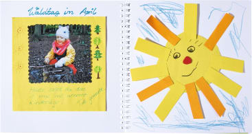Ein aufgeschlagenes Sammelalbum zeigt auf der linken Seite ein Foto eines im Freien spielenden Kindes mit handgeschriebenem Text und Baumschmuck. Die rechte Seite zeigt eine Sonne aus gelben Papierstreifen mit einem lächelnden Gesicht, umgeben von blauen Buntstiftkritzeleien.